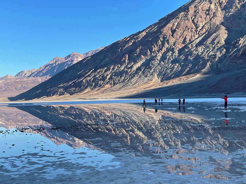A brown hillside slopes up to the right. It is reflected upside-down in calm water. Several people stand in the distance in the water.
Badwater Basin in late December 2023.
NPS