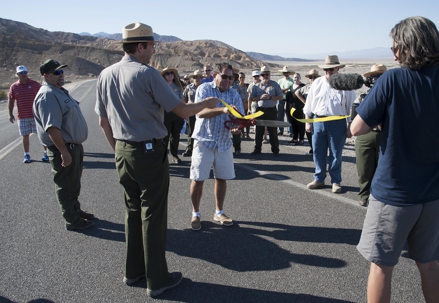 Death Valley National Park Superintendent Mike Reynolds holds a yellow ribbon across Badwater Road while Inyo County Supervisor Matt Kingsley ceremonially cuts it. Park visitors, Furnace Creek Resort staff, and NPS staff watch as Inyo County Supervisor Matt Kingsley ceremonially cuts a ribbon that Park Superintendent Mike Reynolds holds. NPS photo by Birgitta Jansen